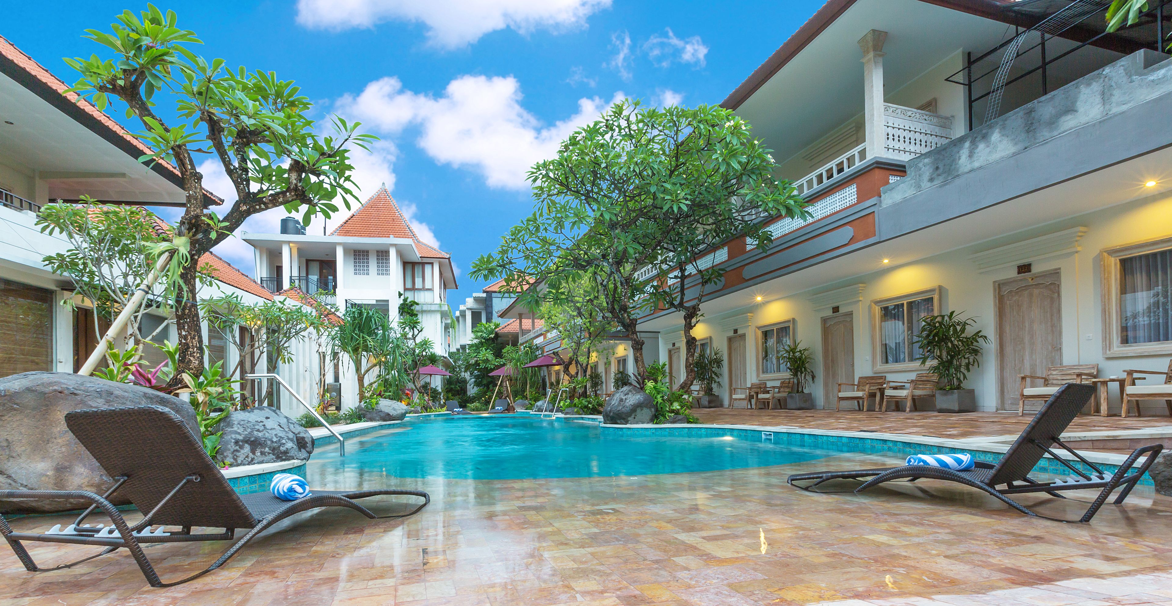 A swimming pool with lounge chairs, surrounded by tropical plants and a building with a tiled roof.