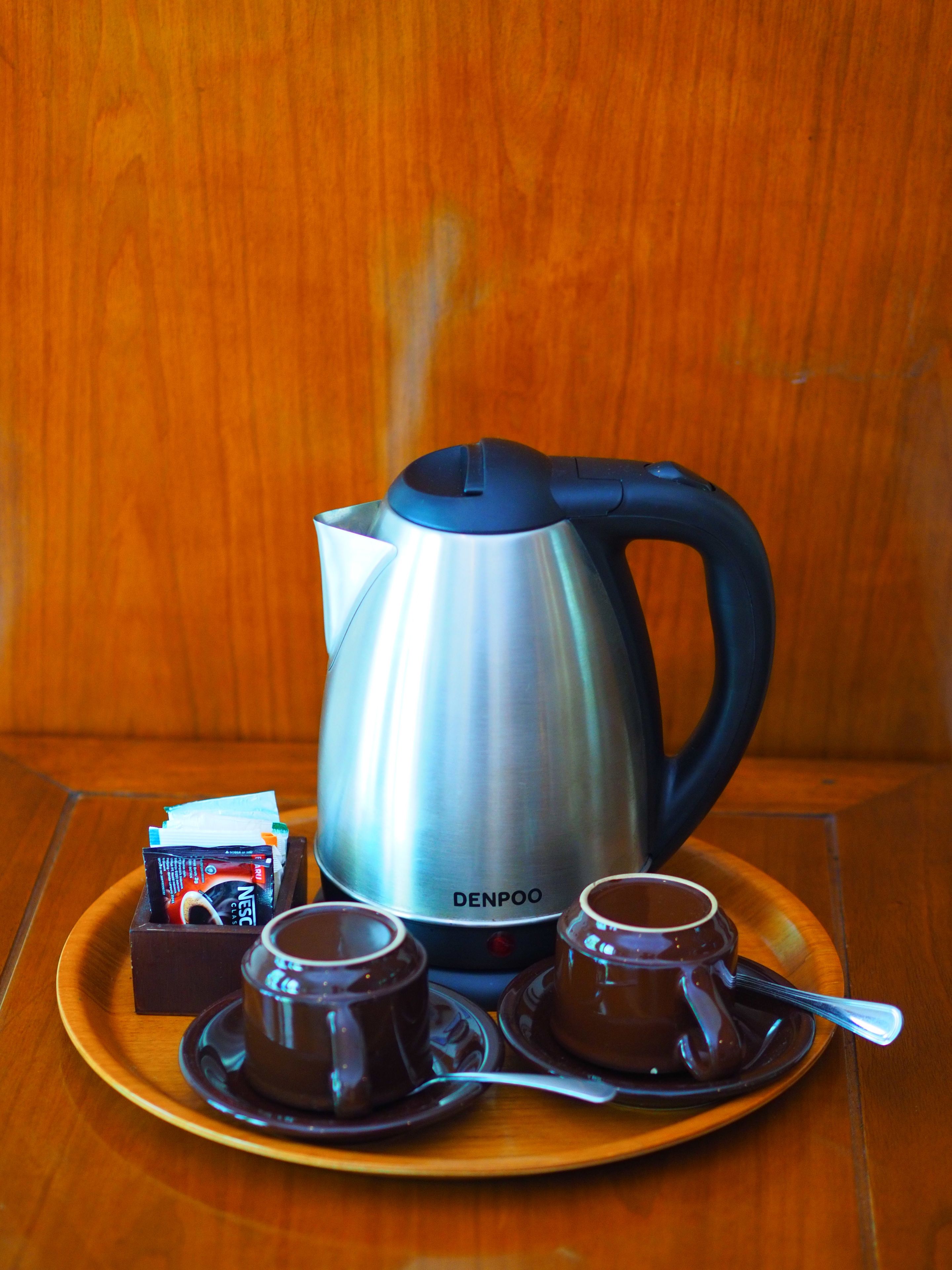 Une bouilloire électrique DENPOO en acier inoxydable, posée sur une table en bois, avec deux tasses marron et une boîte de café instantané Nescafé.