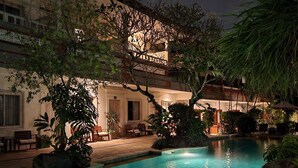 Vista nocturna de un hotel con piscina, mobiliario exterior y abundante vegetación.