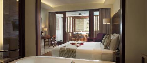 A modern hotel room with a large bathtub filled with flower petals, a bed with a decorative headboard, and a dining area visible through a glass partition.