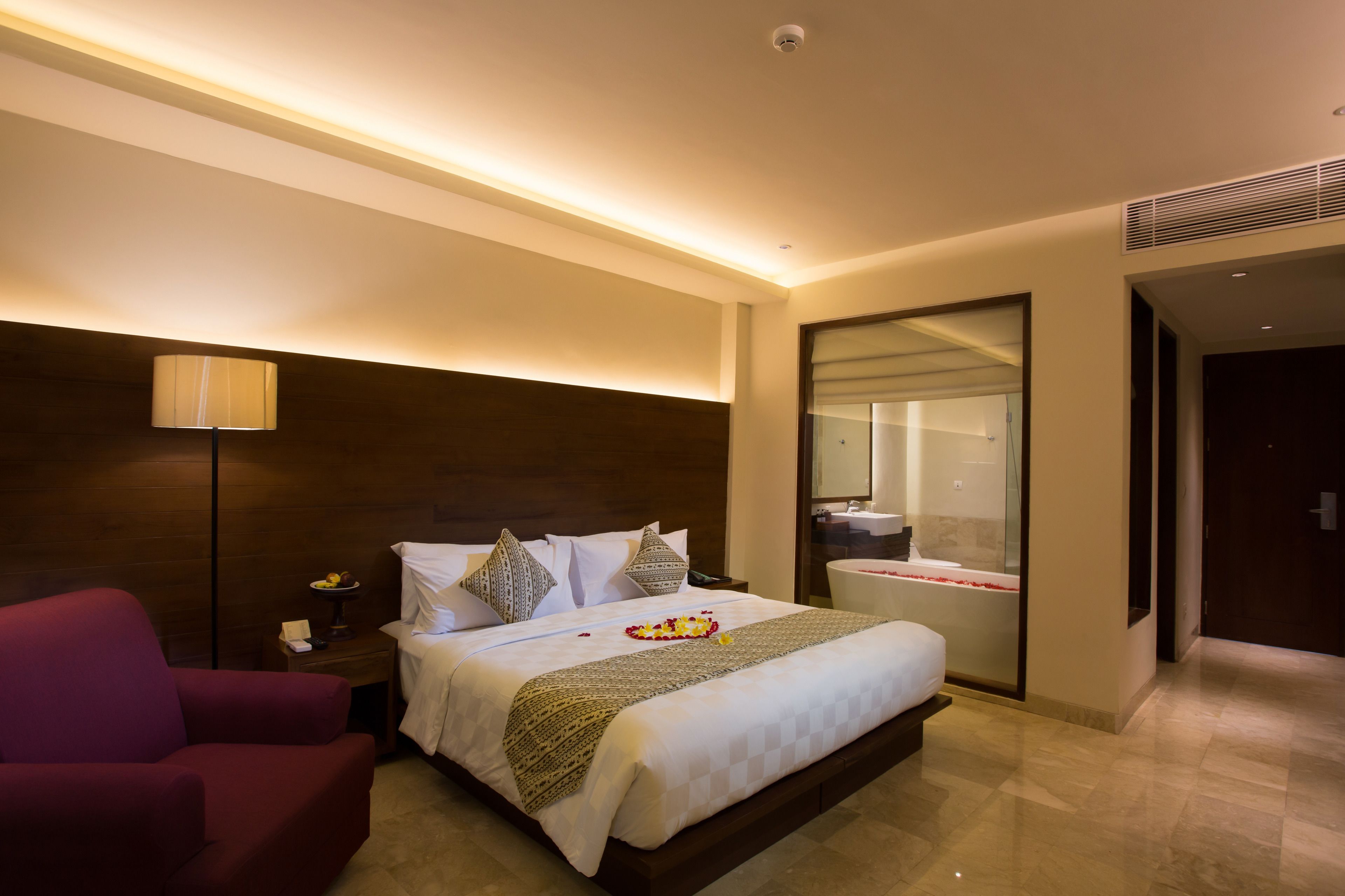 A hotel room with a large bed, a nightstand, a red armchair, a bathroom with a bathtub, and a mirror.