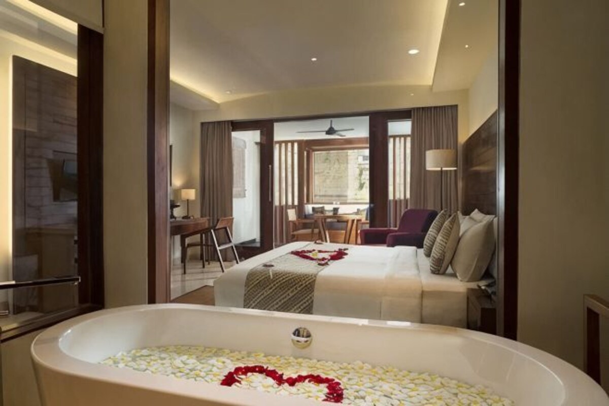 A hotel room with a large bathtub filled with white and red bath products, a bed with a white and gray bedspread, and a dining area with a table and chairs.