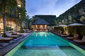 A swimming pool with lounge chairs and umbrellas, surrounded by palm trees and a building with a thatched roof.