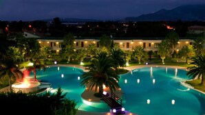 Ein großer, gut beleuchteter Pool mit einer Fontäne, umgeben von Palmen und einem Gebäude mit vielen Fenstern.
