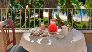 Ein Tisch ist reichhaltig mit Frühstücksleckereien wie Gebäck, Früchten und einem roten Getränk gedeckt, mit Blick auf einen Strand mit Palmen.