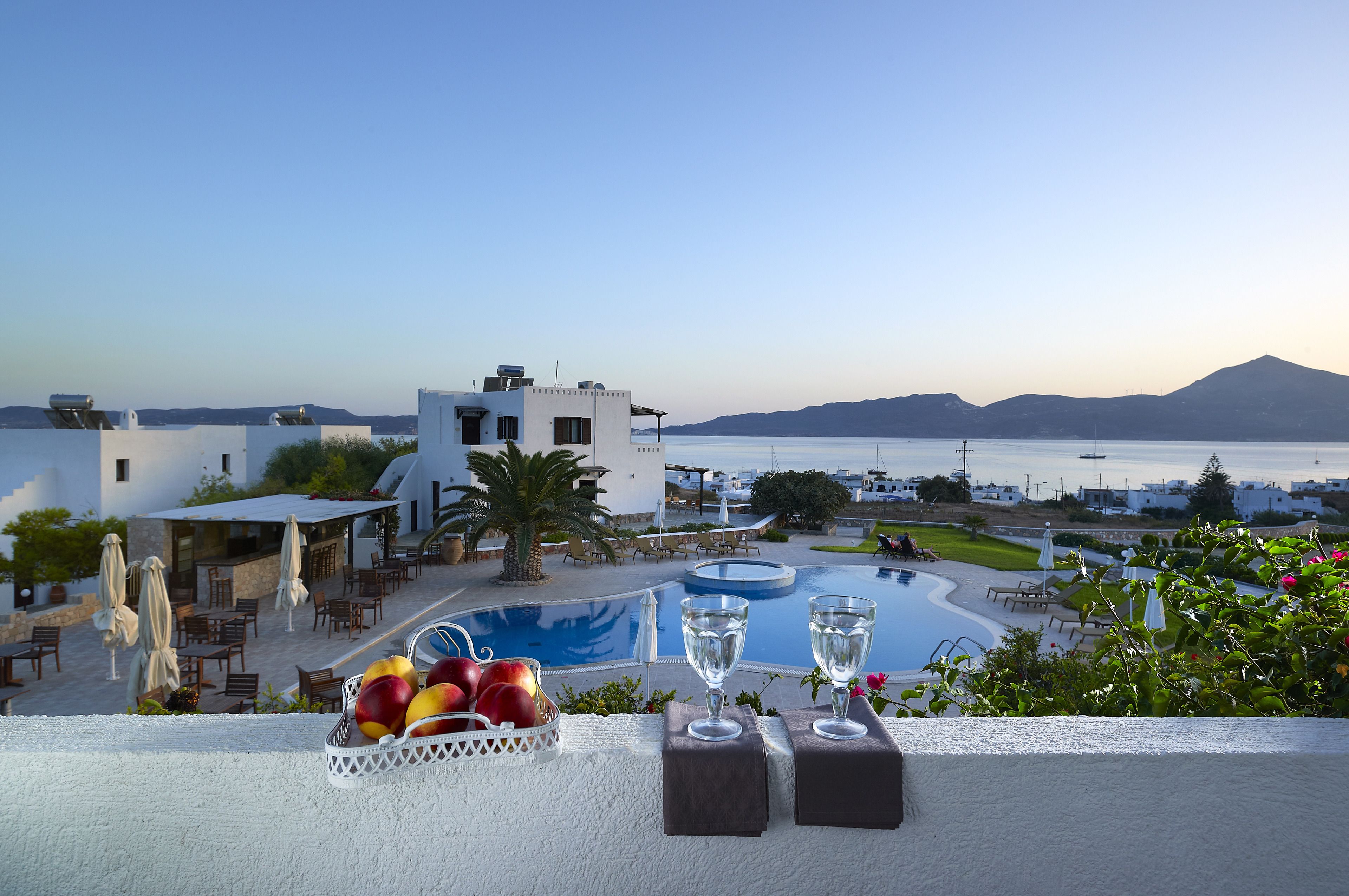 A poolside view with a fruit basket, two glasses, and a table set against a backdrop of a clear sky, a pool, and a mountainous landscape.
