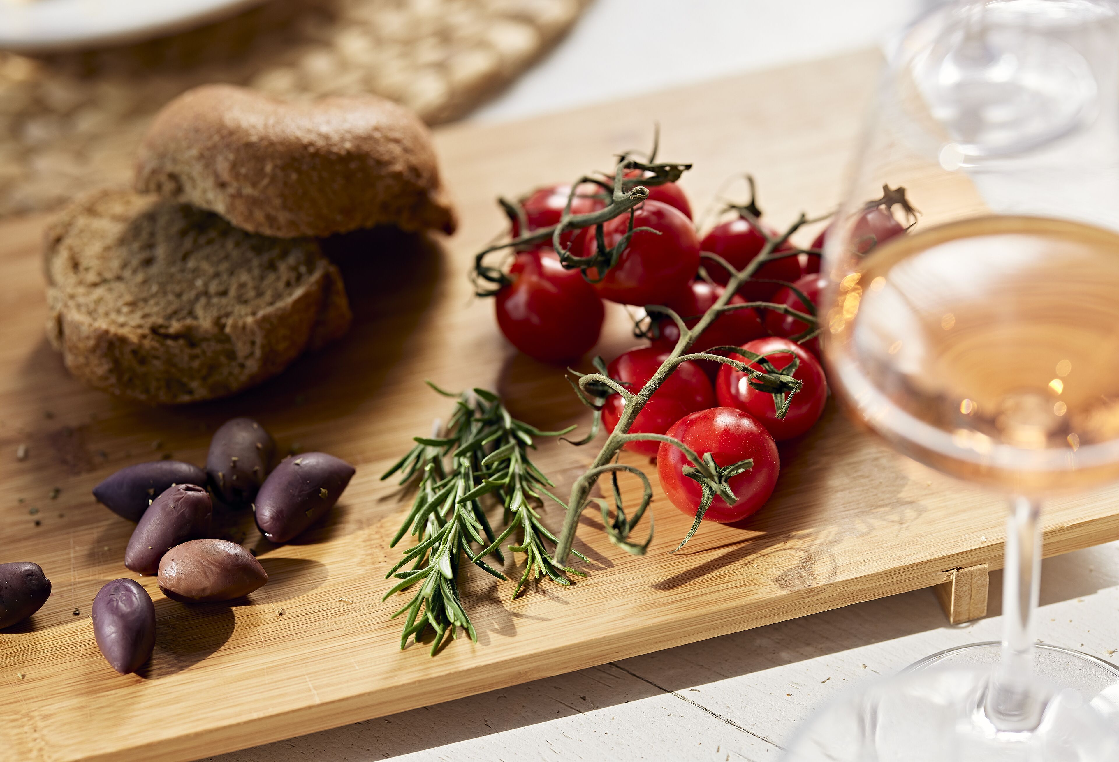 A wooden cutting board with sliced bread, cherry tomatoes, rosemary, and olives.