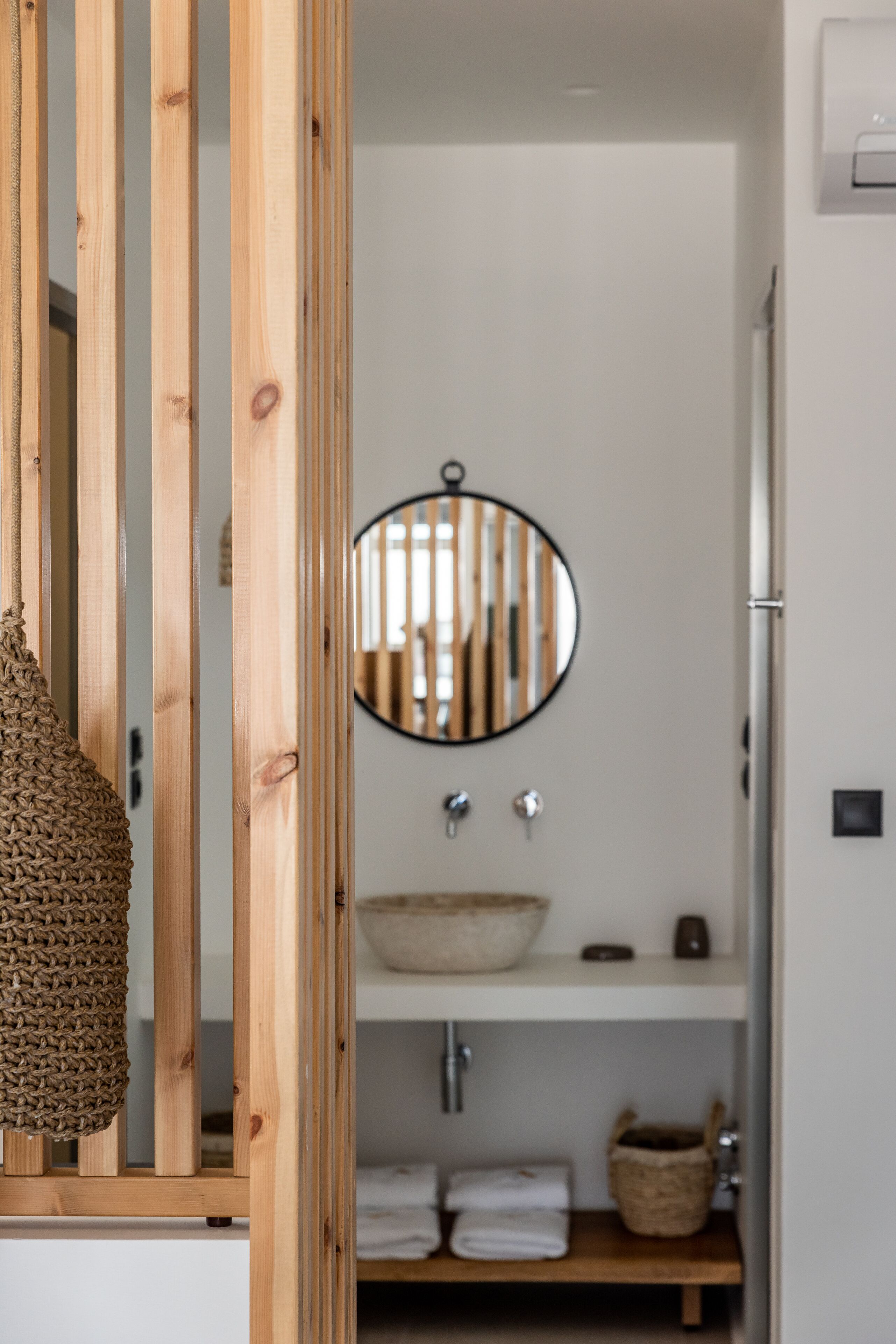 A bathroom with a wooden partition, a round mirror, a white sink, and a shelf with towels and a basket.