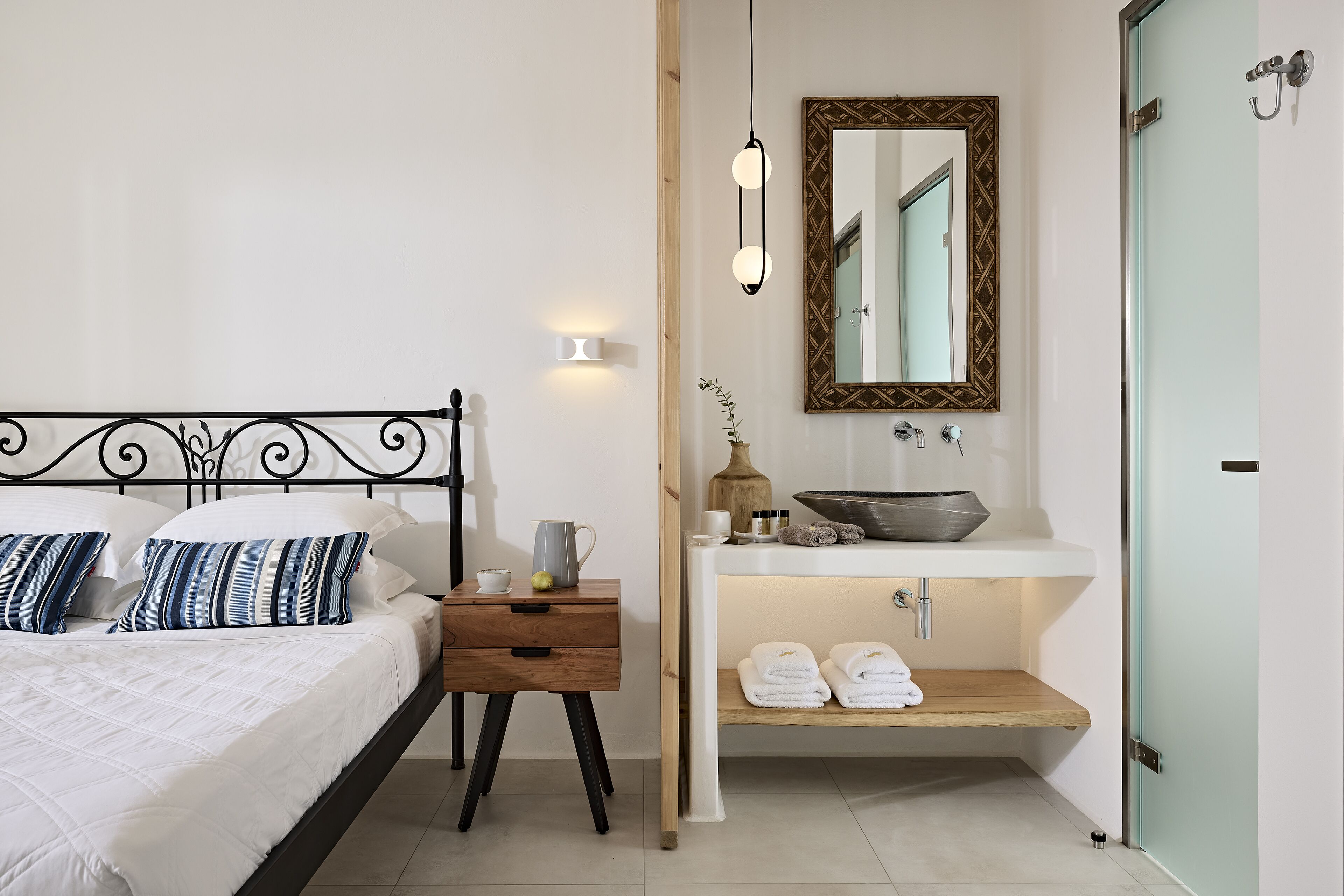 A bedroom with a bed, bedside table, nightstand, and a built-in bathroom with a sink, mirror, and towel rack.