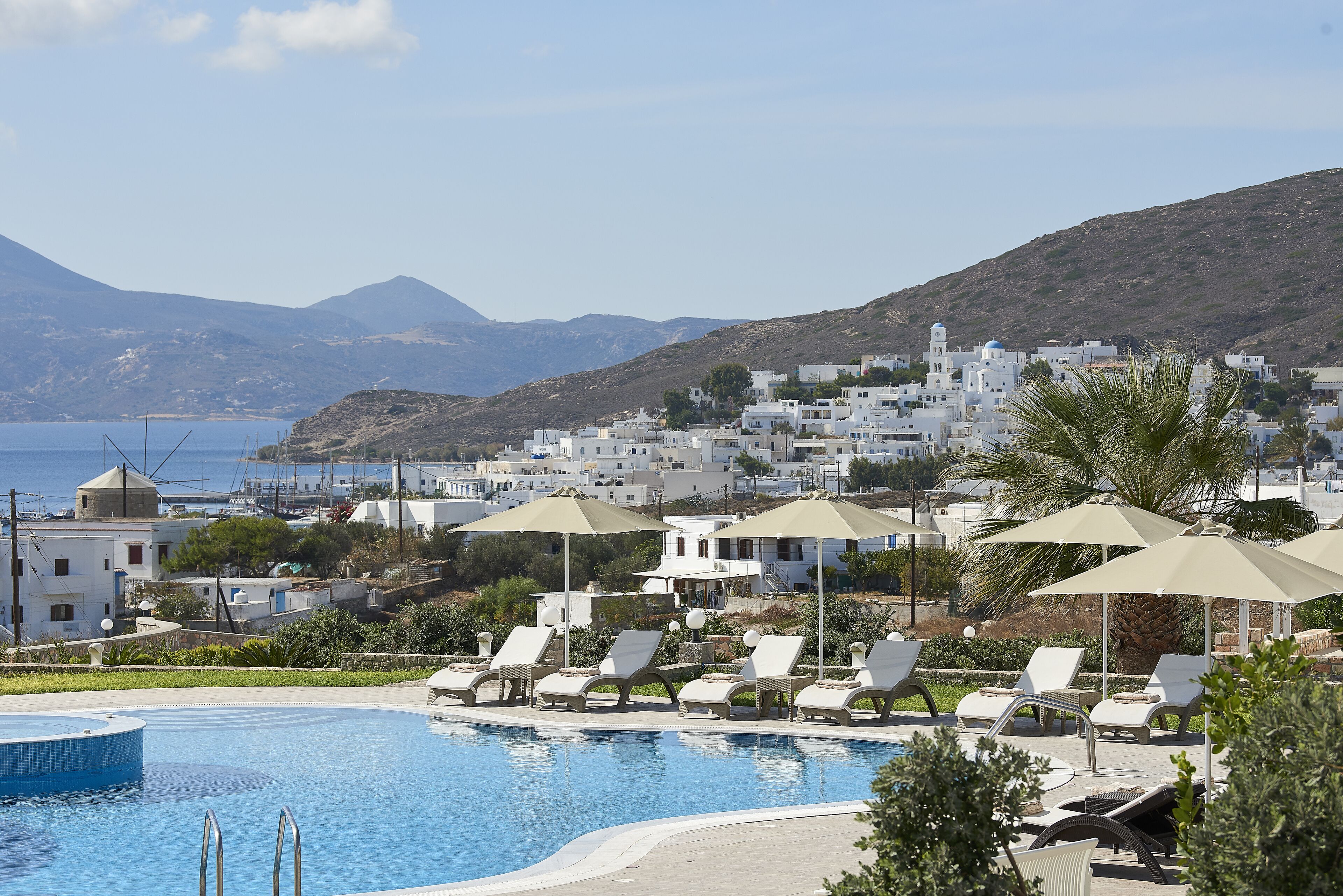 A pool area with lounge chairs and umbrellas, a clear view of the sea, and a town in the background.