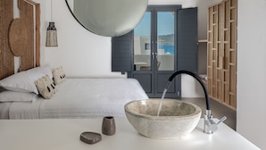 Un dormitorio moderno con una cama grande, un lavabo de piedra con grifo, una lámpara colgante y vistas al mar a través de una puerta.