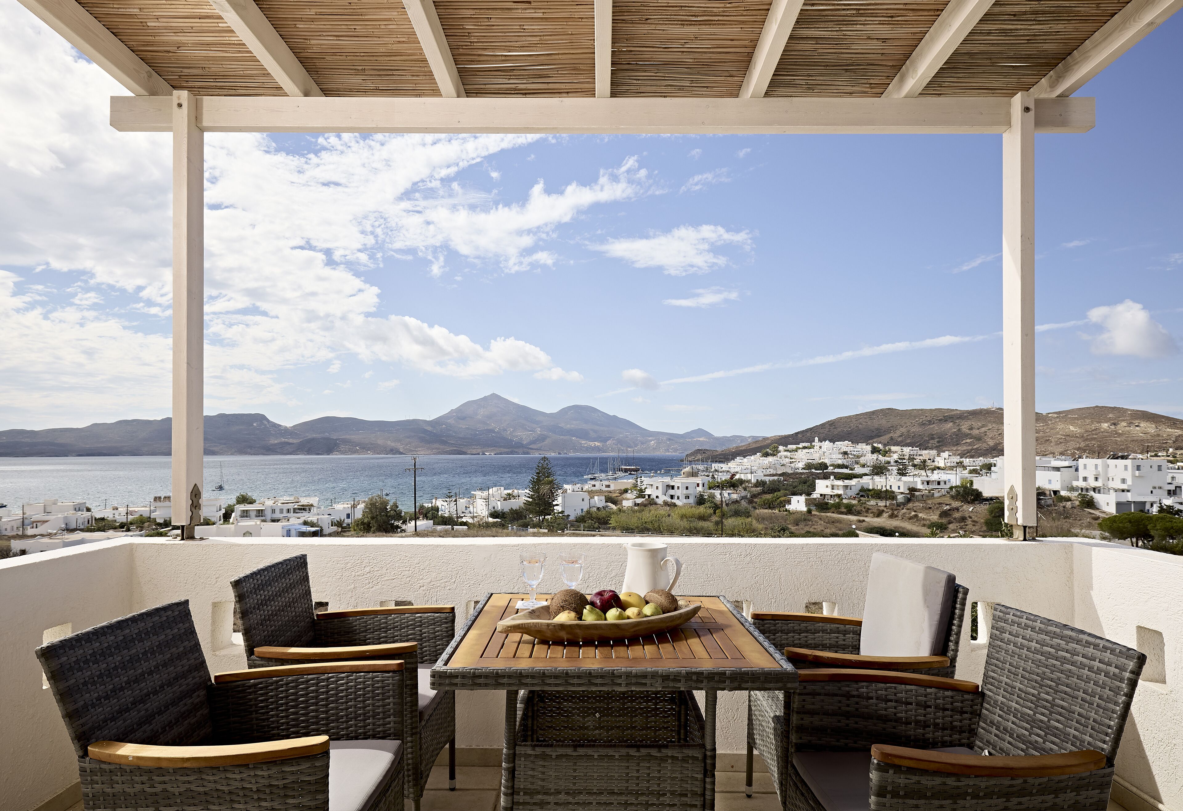 A terrace with wicker chairs, a wooden table, and a fruit bowl. A white pitcher and two glasses are on the table. The terrace overlooks a coastal town with white buildings and mountains in the distance.