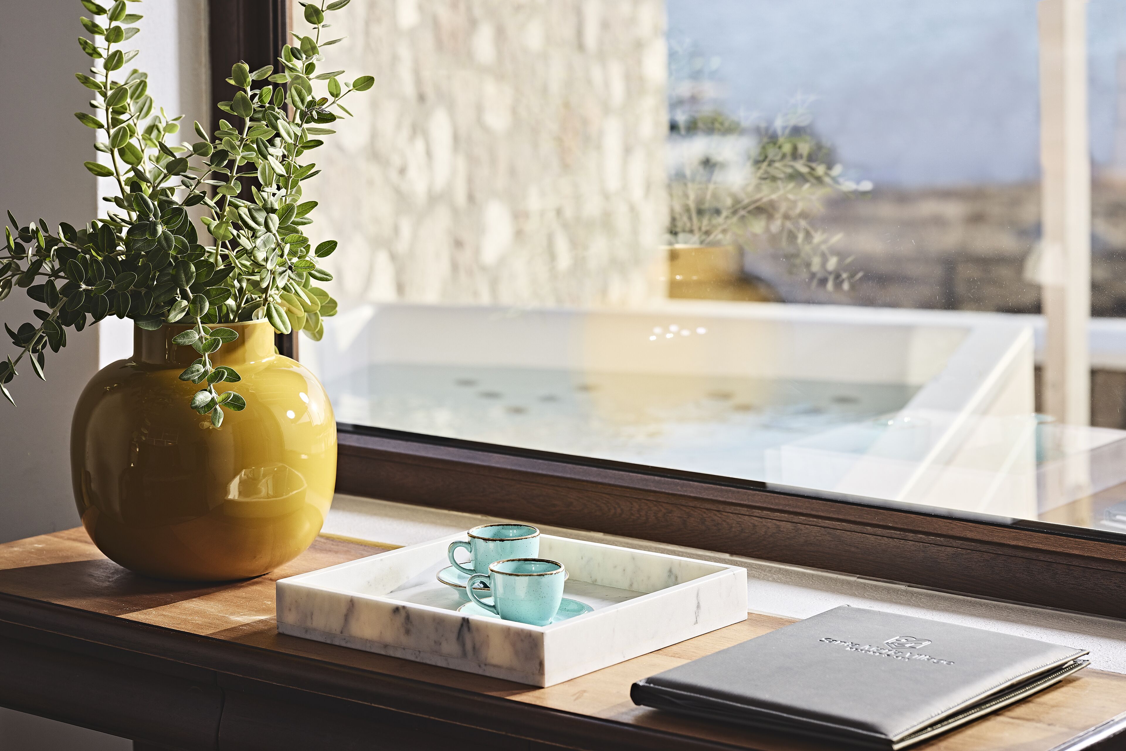 Una ventana con vista a una piscina, un florero amarillo con plantas verdes y una bandeja con dos tazas azules y un libro.