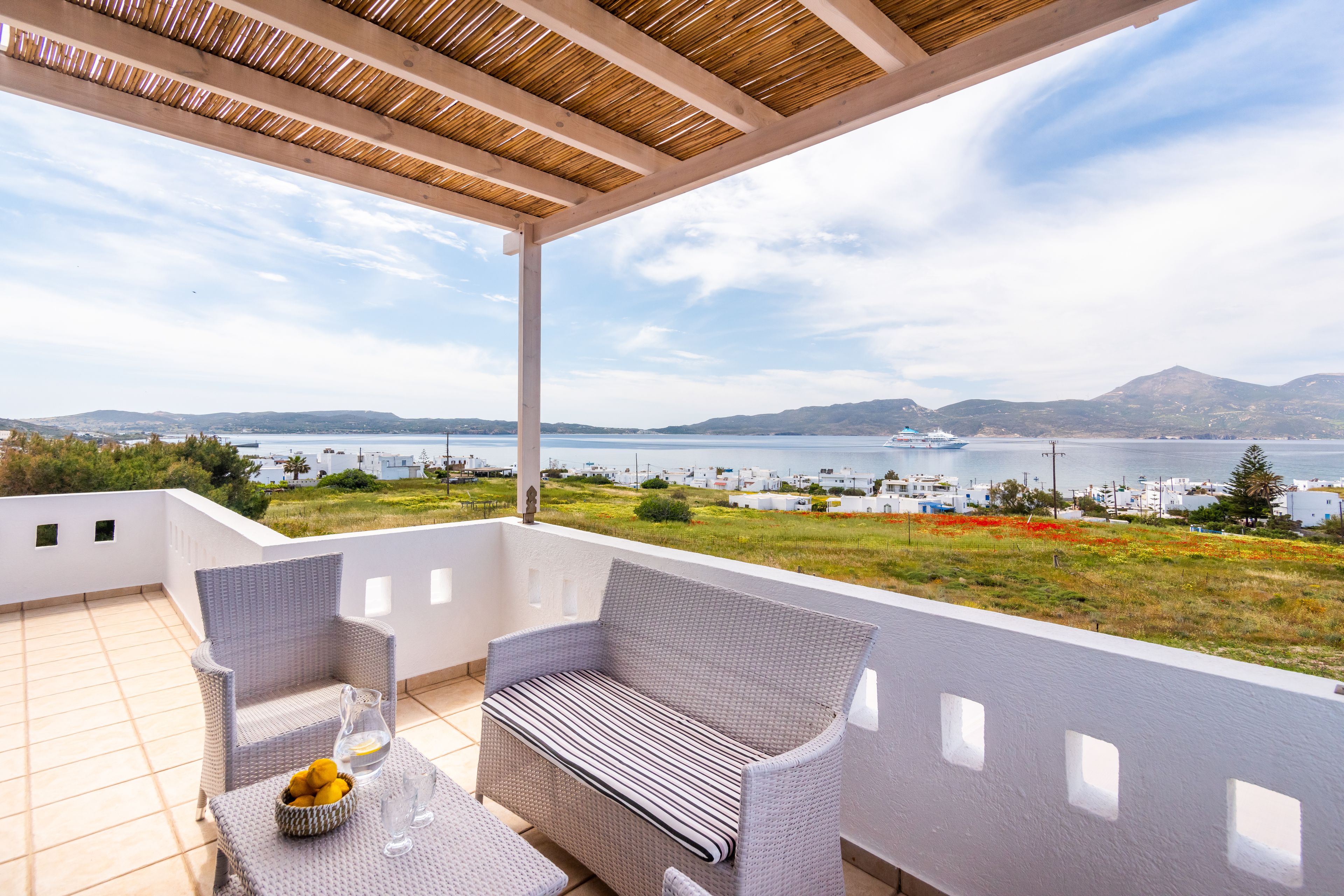 Ein Balkon mit Korbmöbeln, einem Tisch mit Obstschale und Blick auf das Meer und entfernte Gebäude.