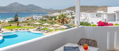 A balcony with a pool, lounge chairs, a table with a book and a glass of drink, and a view of the sea and white buildings.