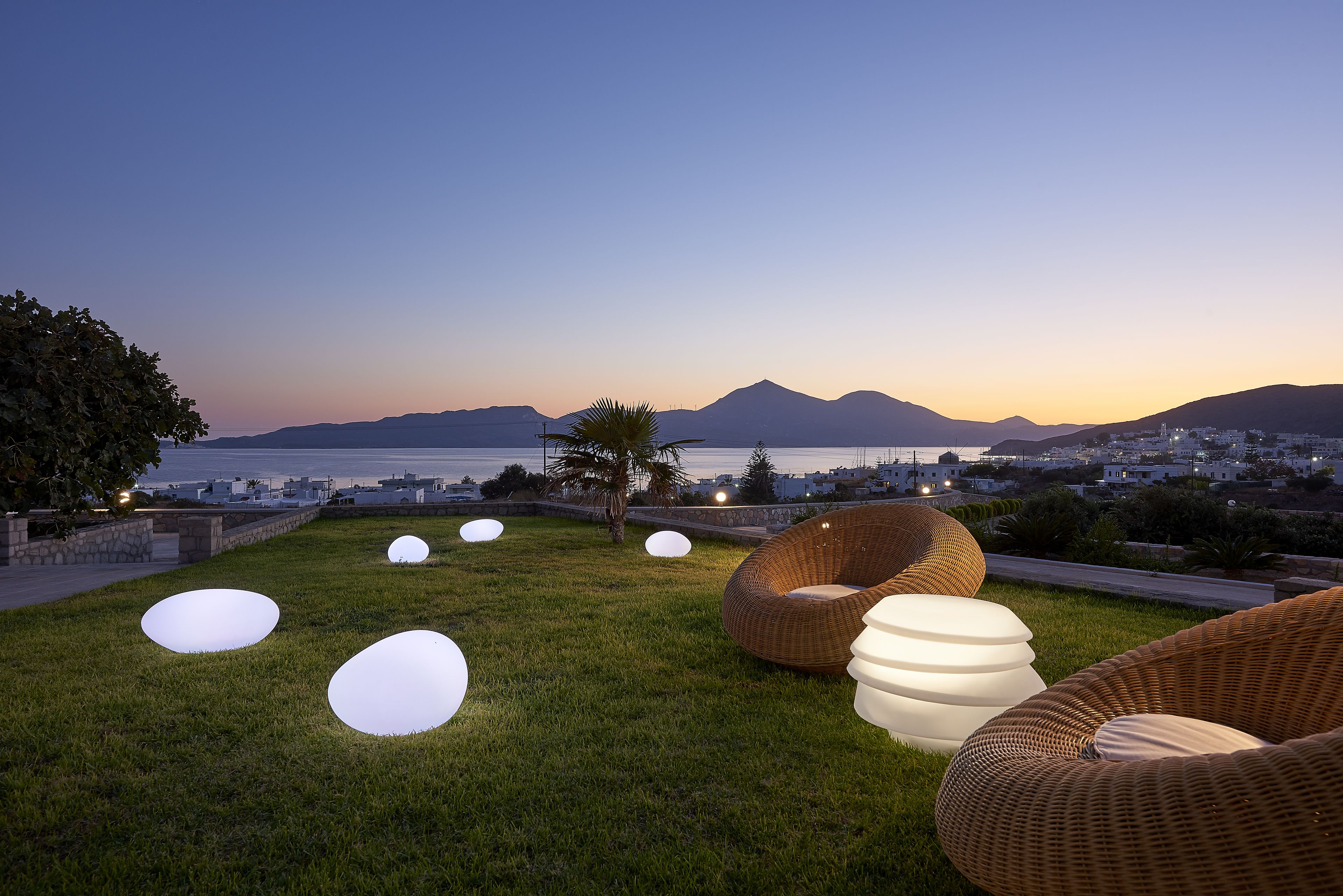 A grassy outdoor area with wicker furniture and illuminated spherical objects, overlooking a coastal cityscape with mountains in the background.