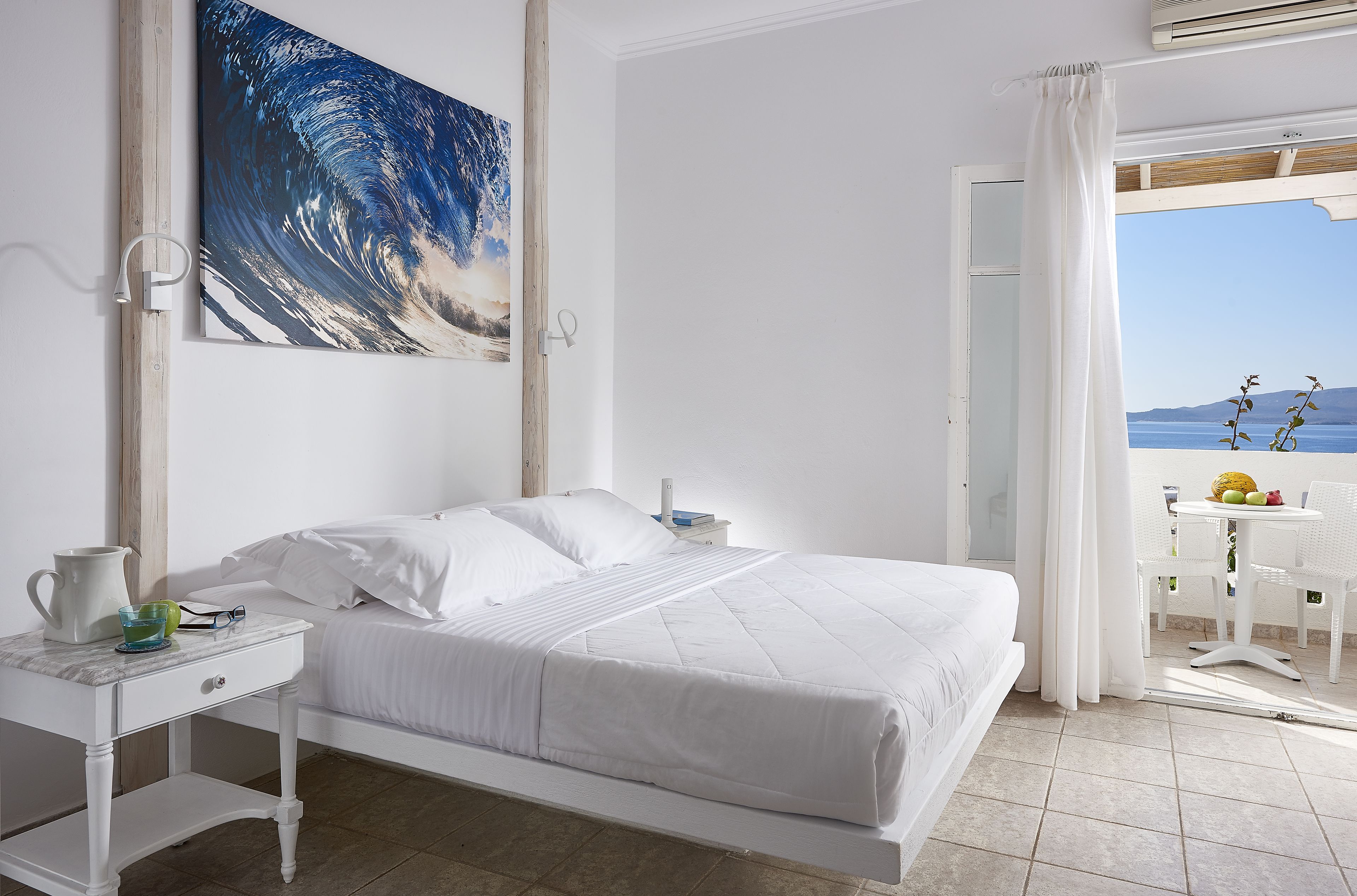 Une chambre à coucher avec un lit, une table de chevet, un tableau et une vue sur la mer par la fenêtre.