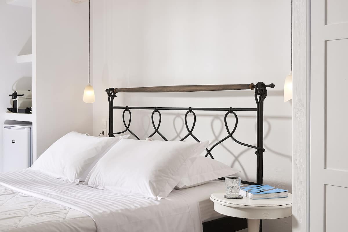 Ein ordentlich bezogenes Bett mit weißen Leinen, ein Nachttisch mit Glas und Notizbuch sowie eine wandmontierte Lampe.
