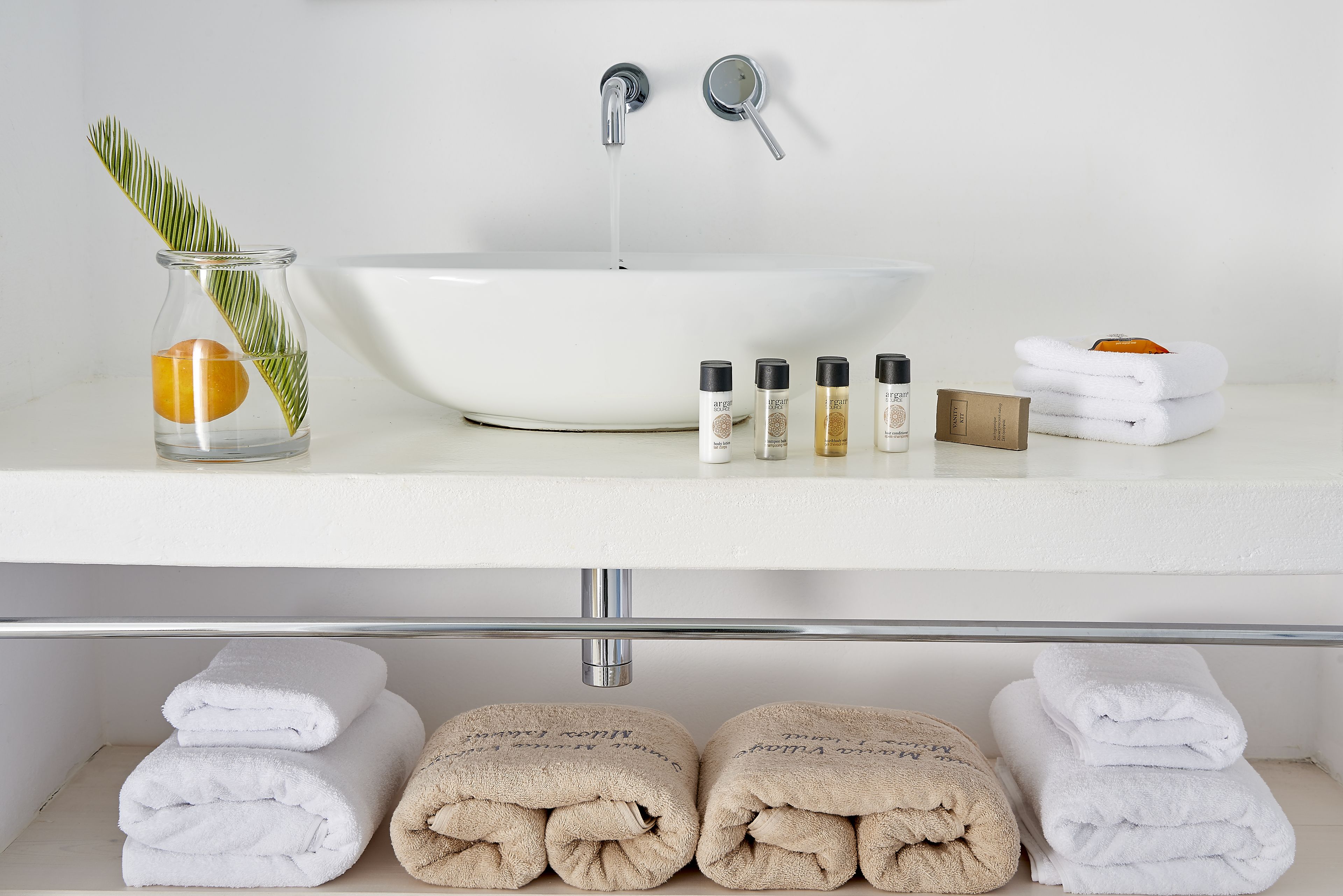 A bathroom with a white sink, two faucets, a glass vase with a leaf and an orange, and several bottles of toiletries.
