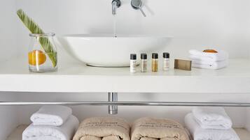 A bathroom with a white sink, two faucets, a glass vase with a leaf and an orange, and several bottles of toiletries.
