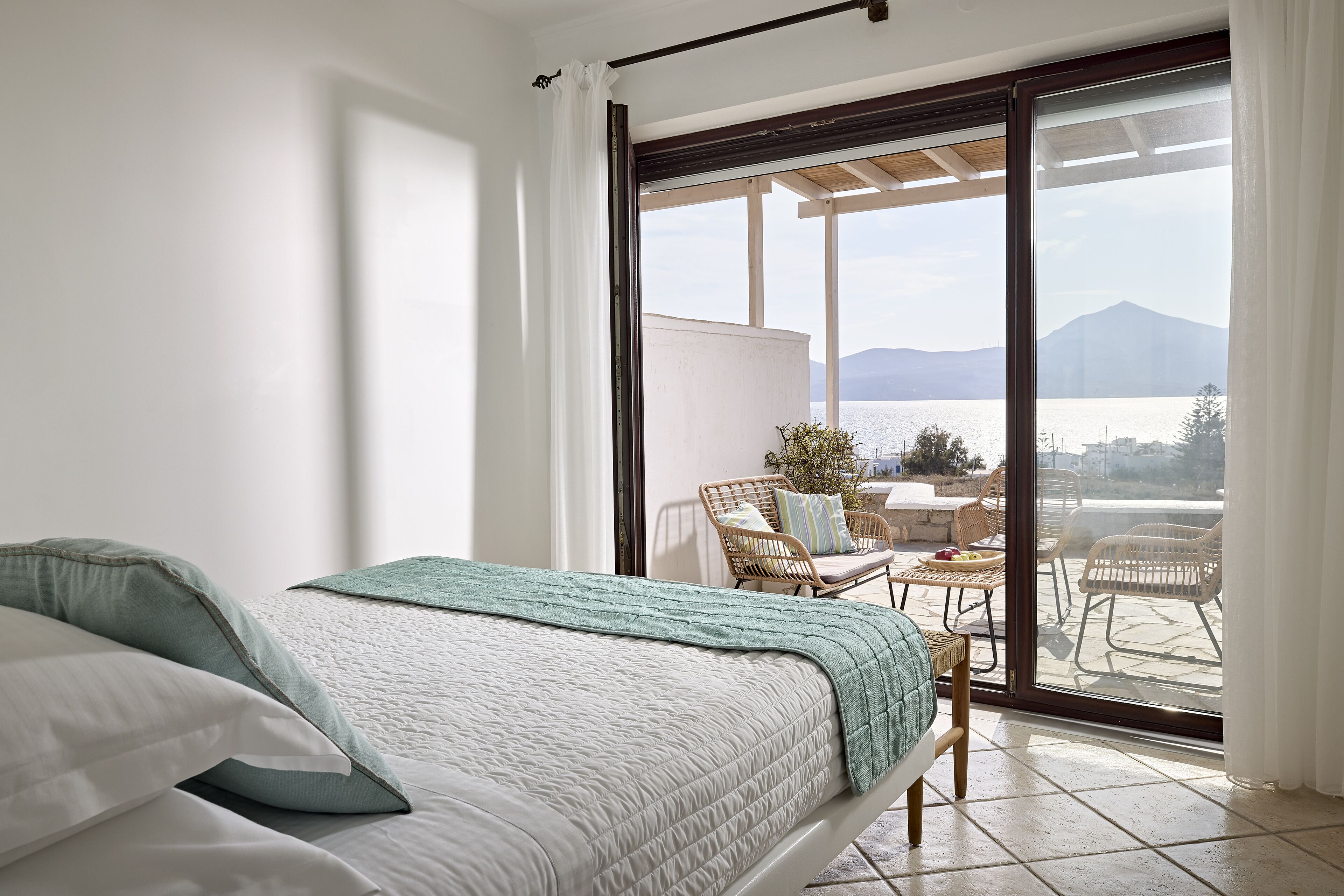 A bedroom with a bed, a view of a balcony with outdoor furniture, and a mountain in the background.
