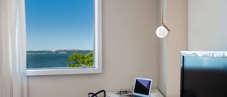 A modern room with a desk, laptop, and TV. A window offers a view of the sea and a distant cityscape.