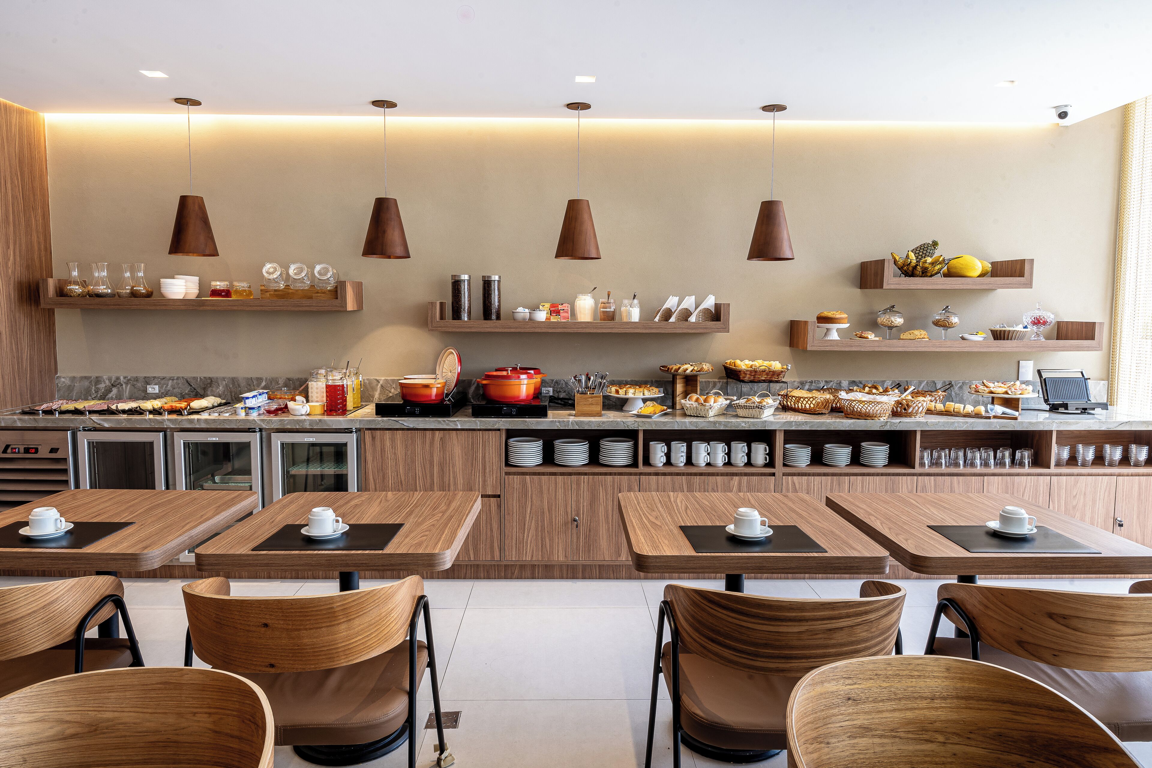 A modern café with wooden furniture, a central counter with various food items, and hanging pendant lights.