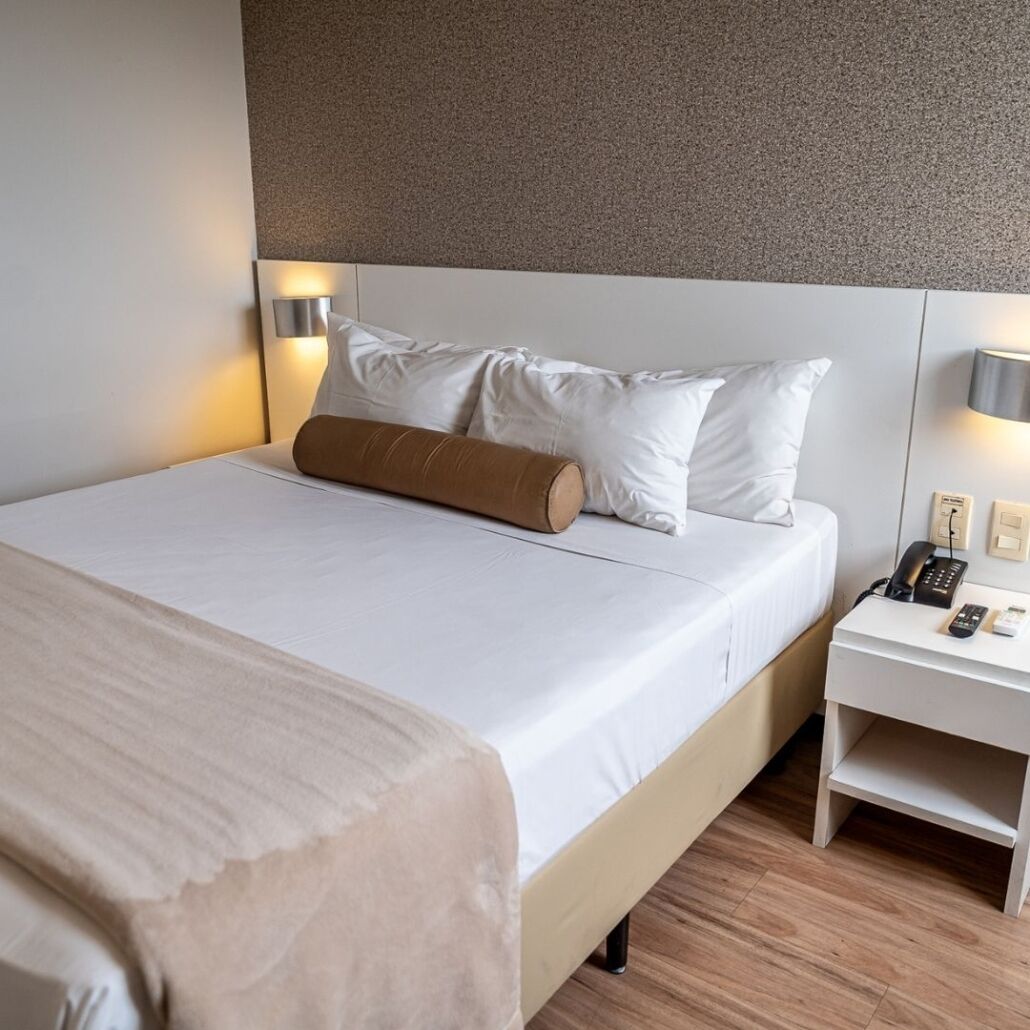 A hotel room with a neatly made bed, a bedside table with a phone and a lamp, and a wall-mounted light fixture.