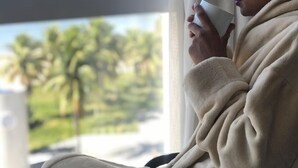 A person in a bathrobe sitting by a window with a view of a body of water and greenery. - Píer Vitória Hotel (Vitoria)