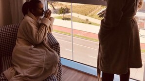 A room with a large window overlooking a beach and water. There is a person sitting on a couch and another standing by the window. - Píer Vitória Hotel (Vitoria)