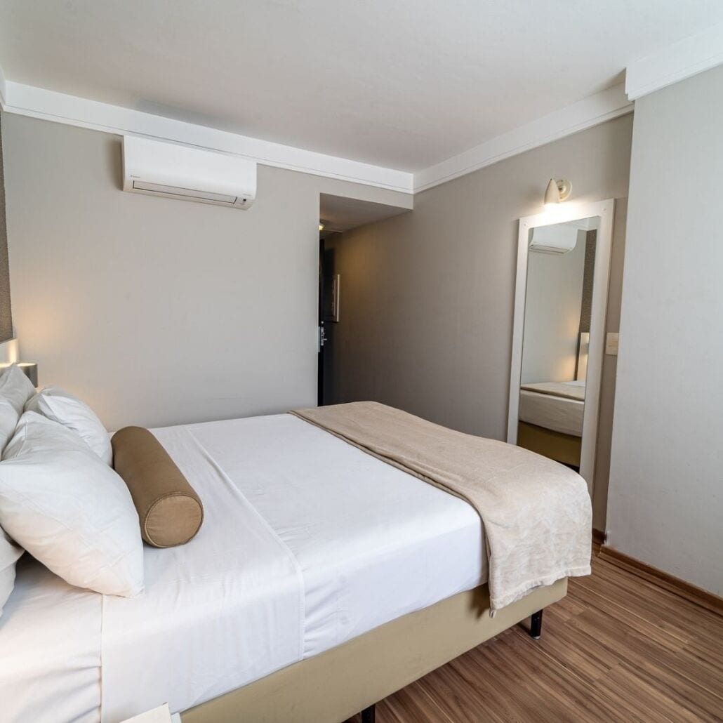 Una cama bien hecha con sábanas blancas y un plaid beige, una lámpara de noche y un aire acondicionado montado en la pared.
