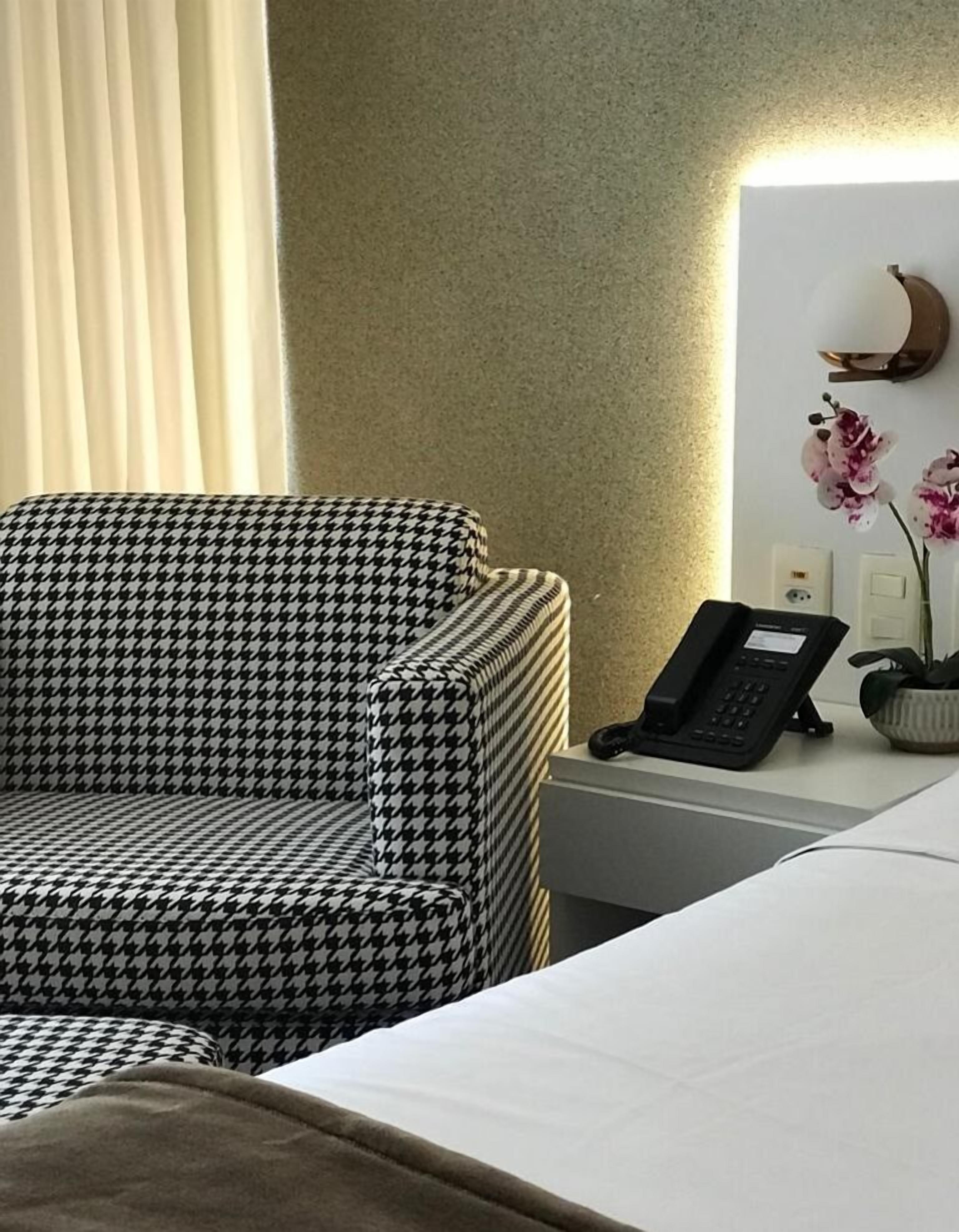 A hotel room with a houndstooth patterned armchair, a bedside telephone, and a vase with flowers on the nightstand.