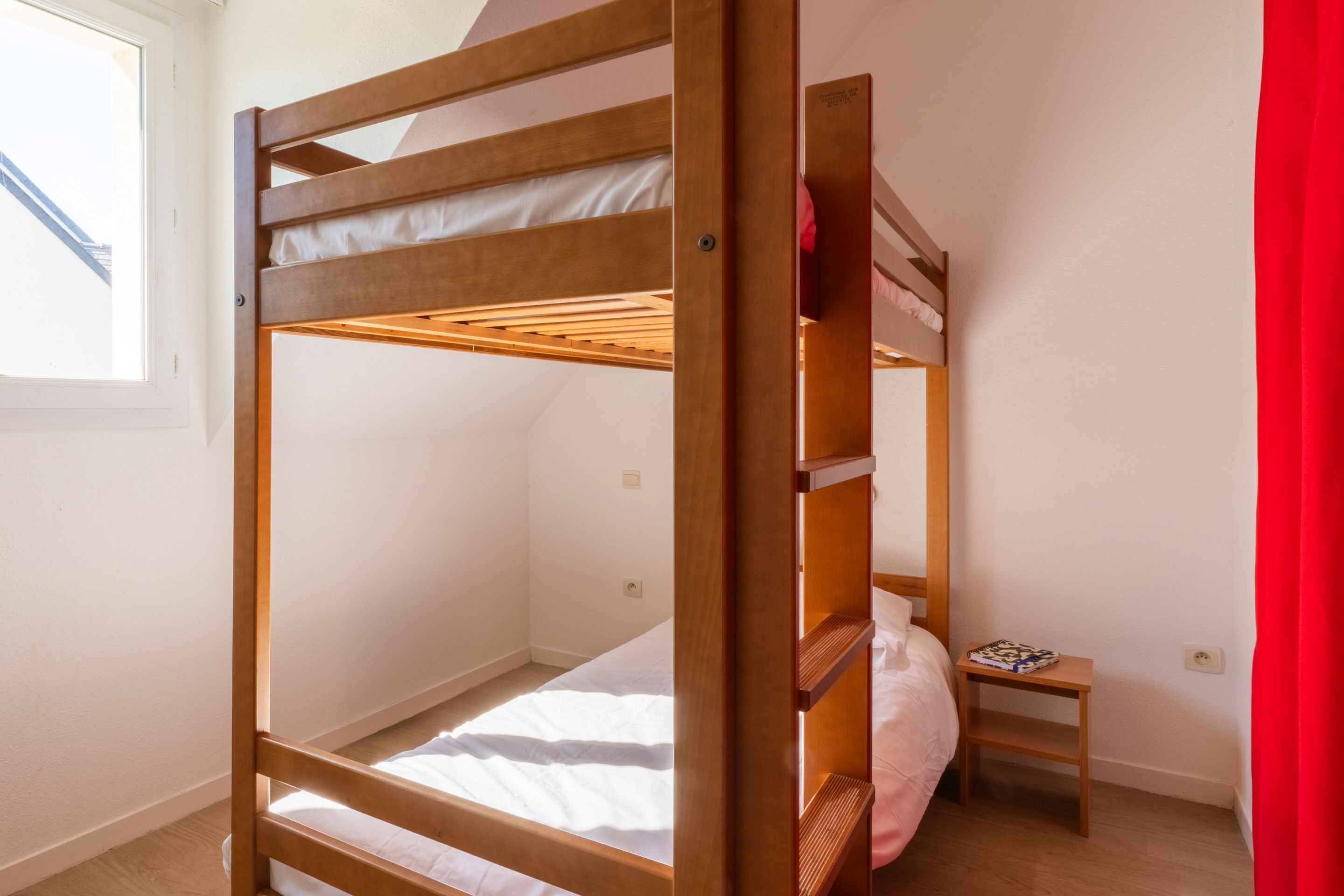 Une chambre avec un lit superposé, une petite table de chevet en bois et une fenêtre.