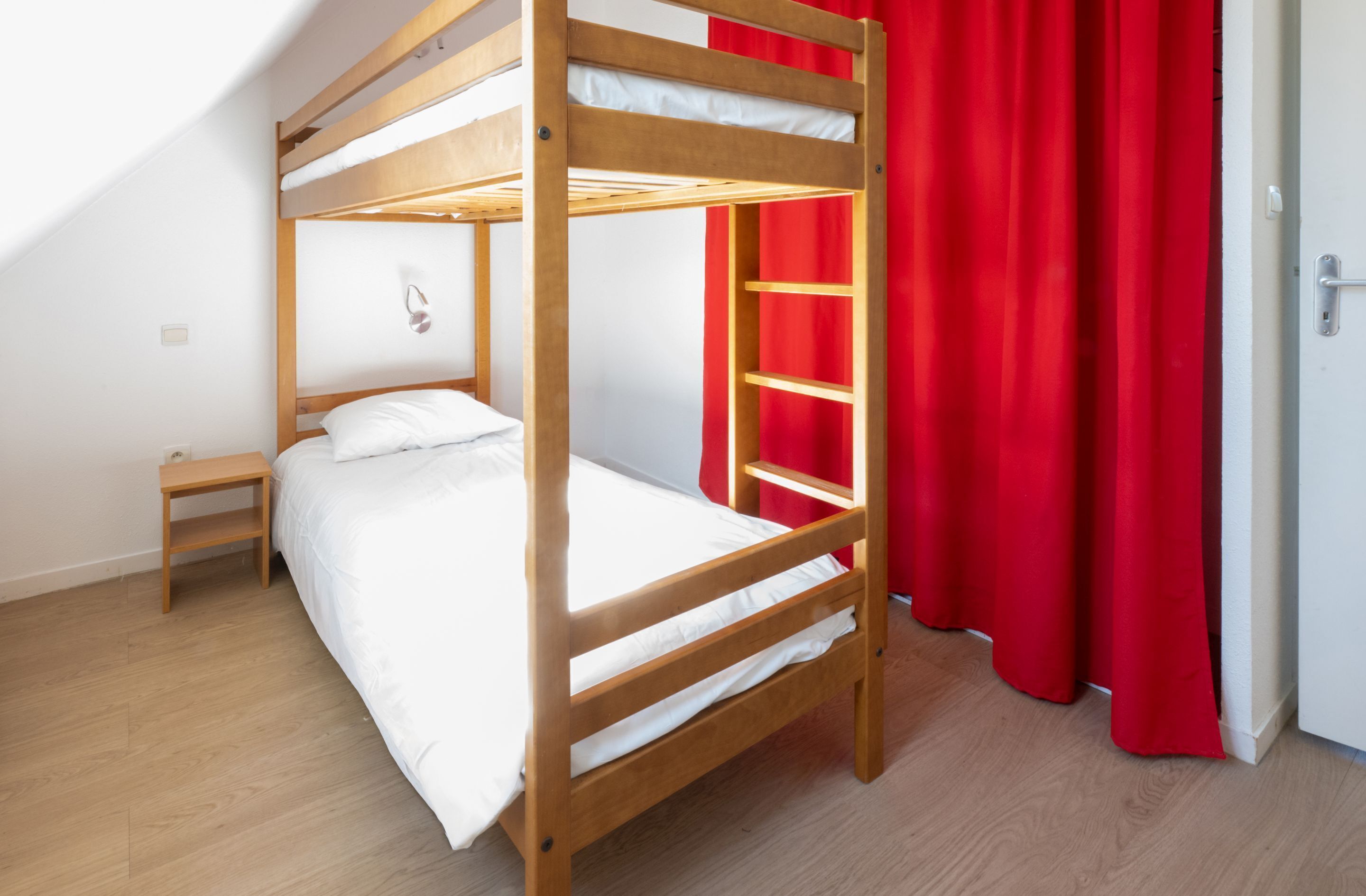 A bunk bed with a wooden frame and white bedding, a small wooden nightstand, and a red curtain.