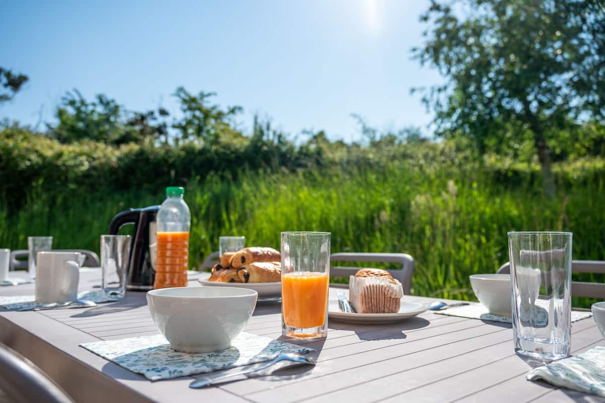 A breakfast table set outdoors with a bottle of orange juice, a glass of orange juice, a bowl, a plate with pastries, and a glass of water.