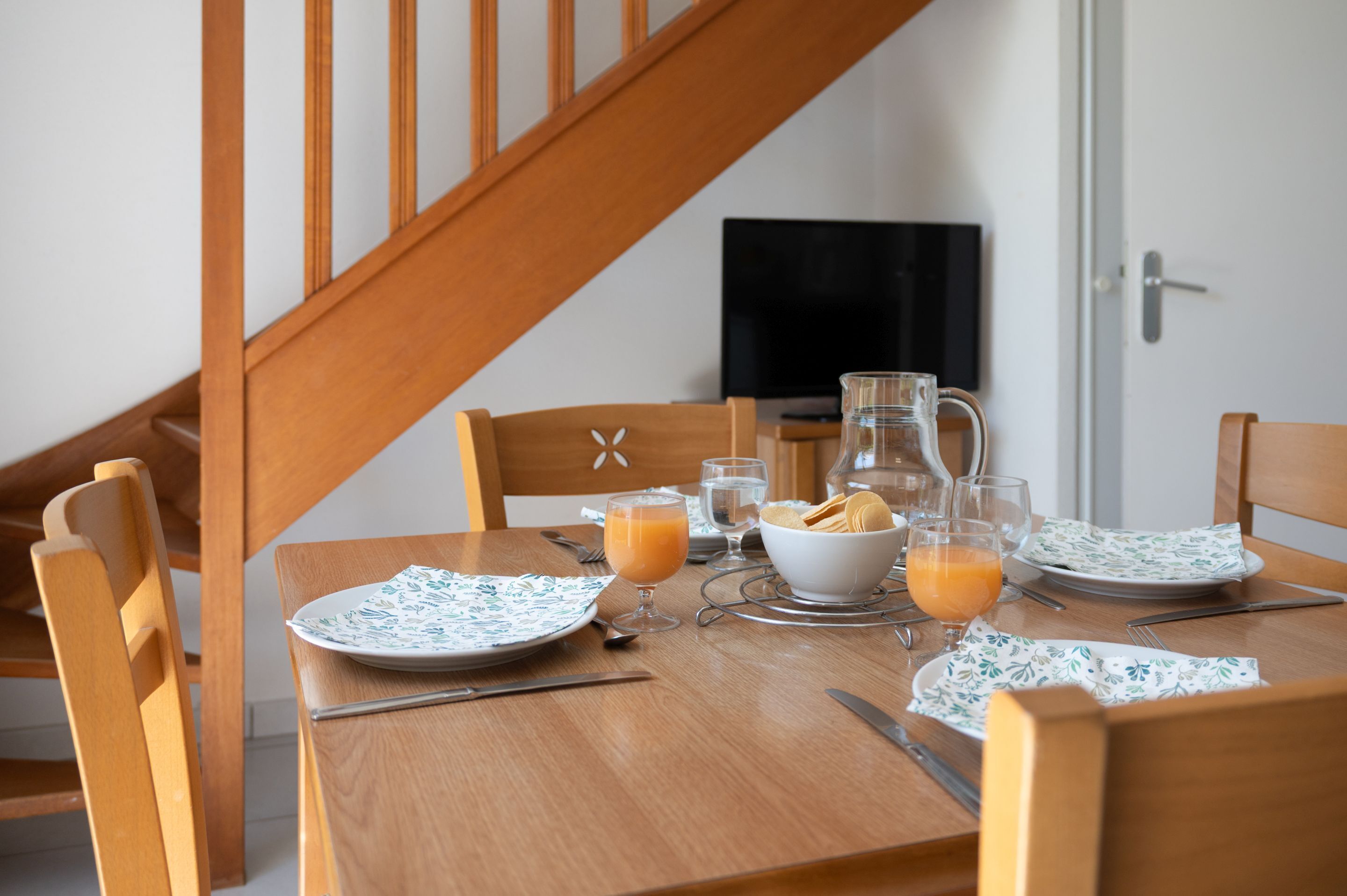 Une table à manger dressée avec des assiettes, des verres et une carafe de jus d’orange.