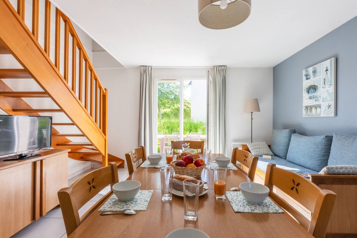 A dining area with a wooden table set for four, surrounded by chairs, a television on a wooden cabinet, and a staircase leading to an upper floor.