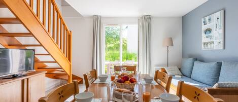 A dining area with a wooden table set for four, surrounded by chairs, a television on a wooden cabinet, and a staircase leading to an upper floor.