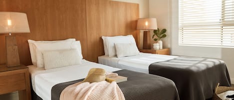 A hotel room with two beds, wooden headboard, bedside lamps, a window with blinds, and a small plant on the nightstand.