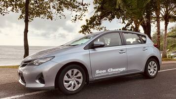 A silver hatchback car parked on a road with a view of the ocean and palm trees.