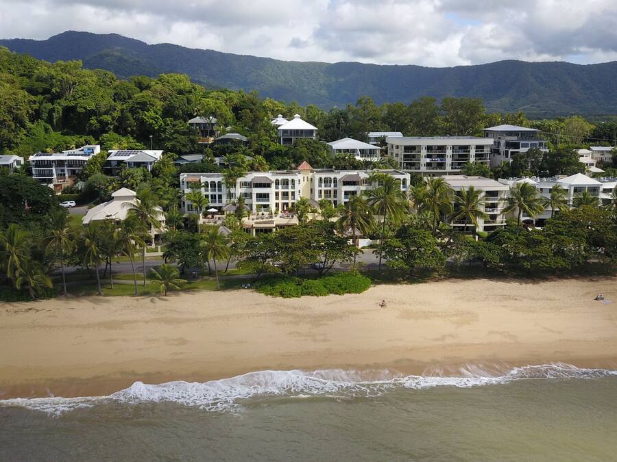 Un hotel frente a la playa con varias edificaciones, una playa de arena y montañas al fondo.