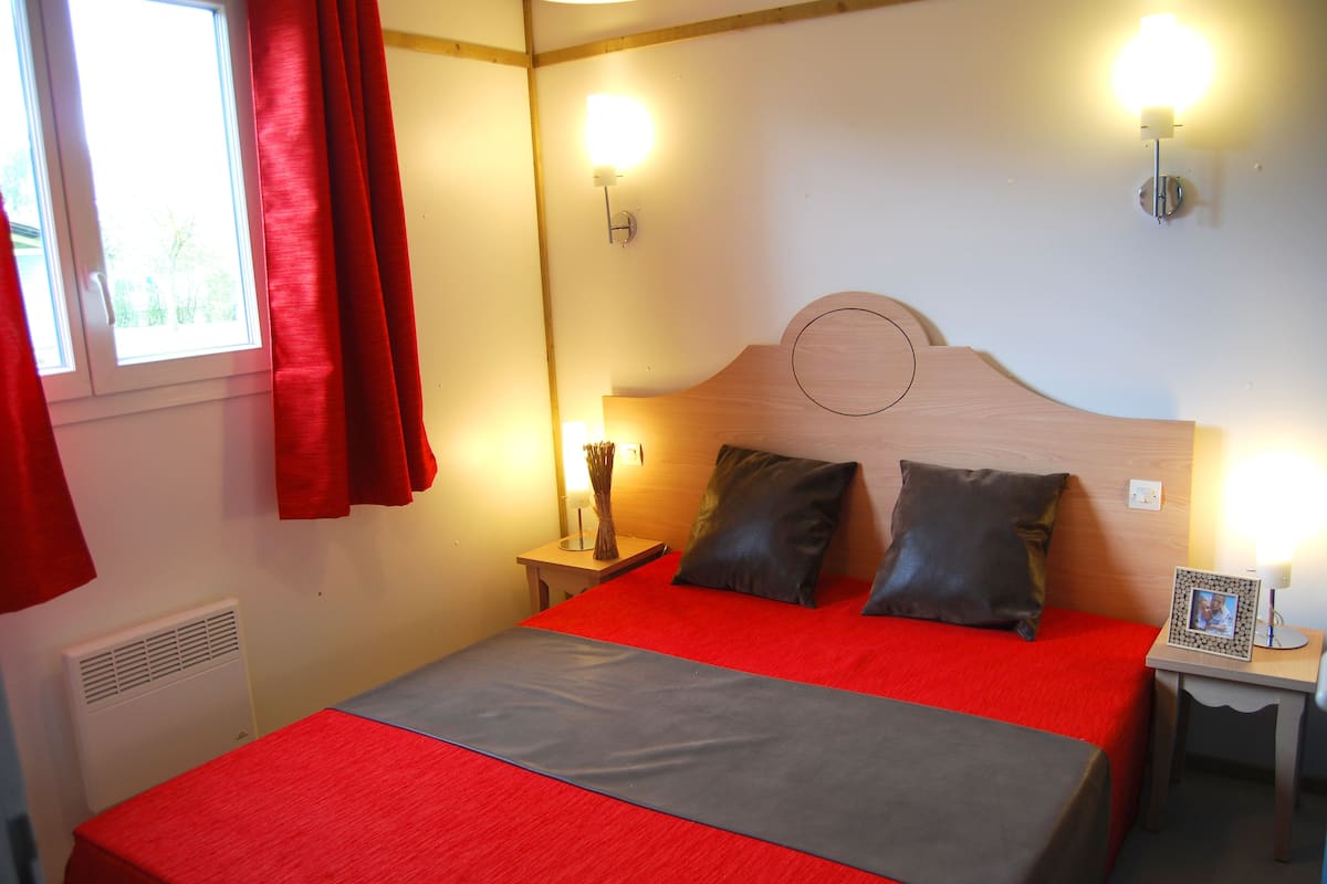 A hotel room with a bed, red curtains, a bedside table with a lamp, and a wall-mounted light fixture.