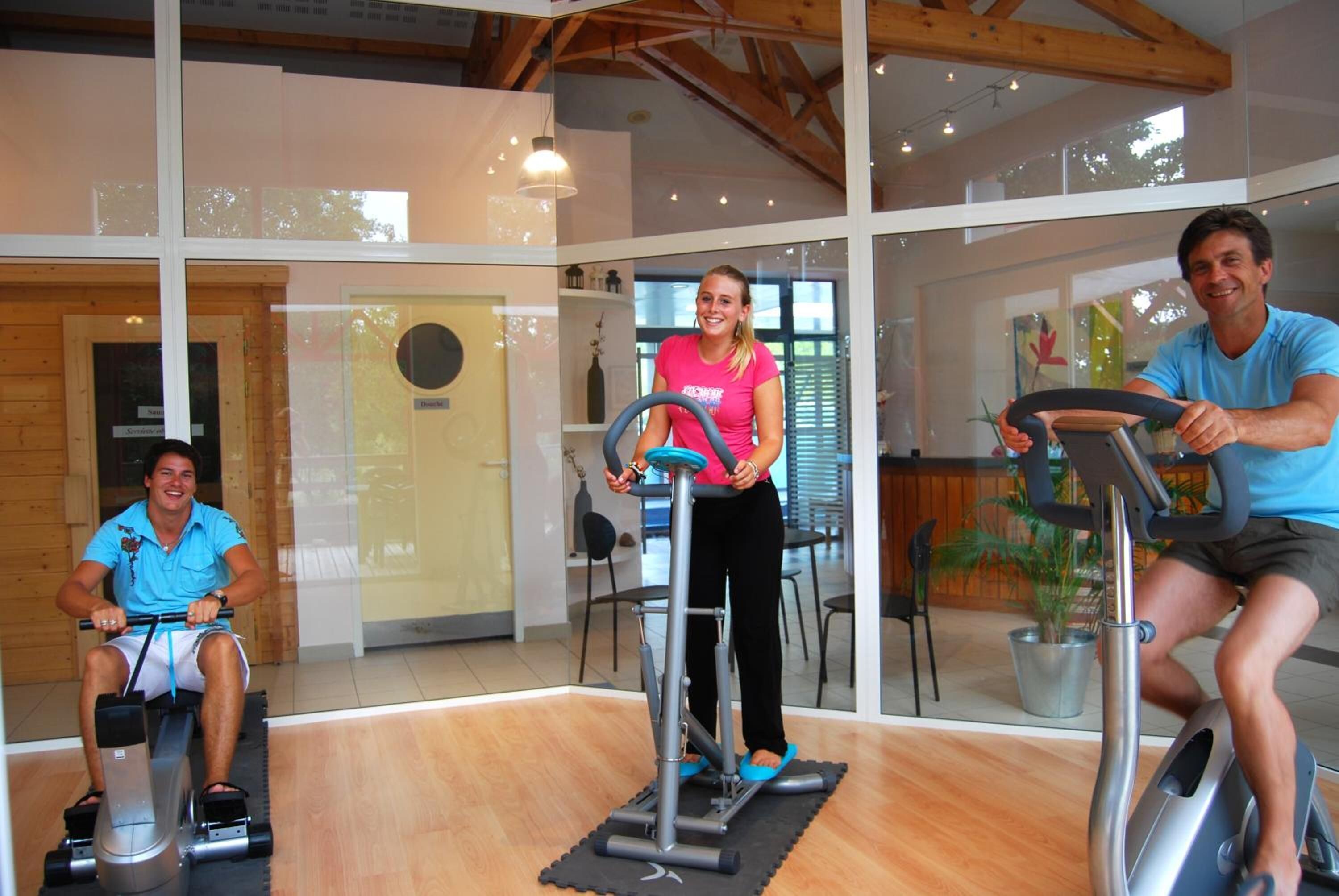Three people using stationary bikes in a well-lit indoor space with wooden beams and large windows.