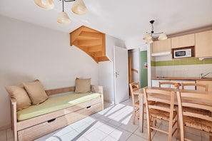 A compact living space with a bed, a dining table with chairs, a kitchen area with cabinets and a microwave, and a staircase. - Résidence Odalys Le Hameau du Lac (Rignac)