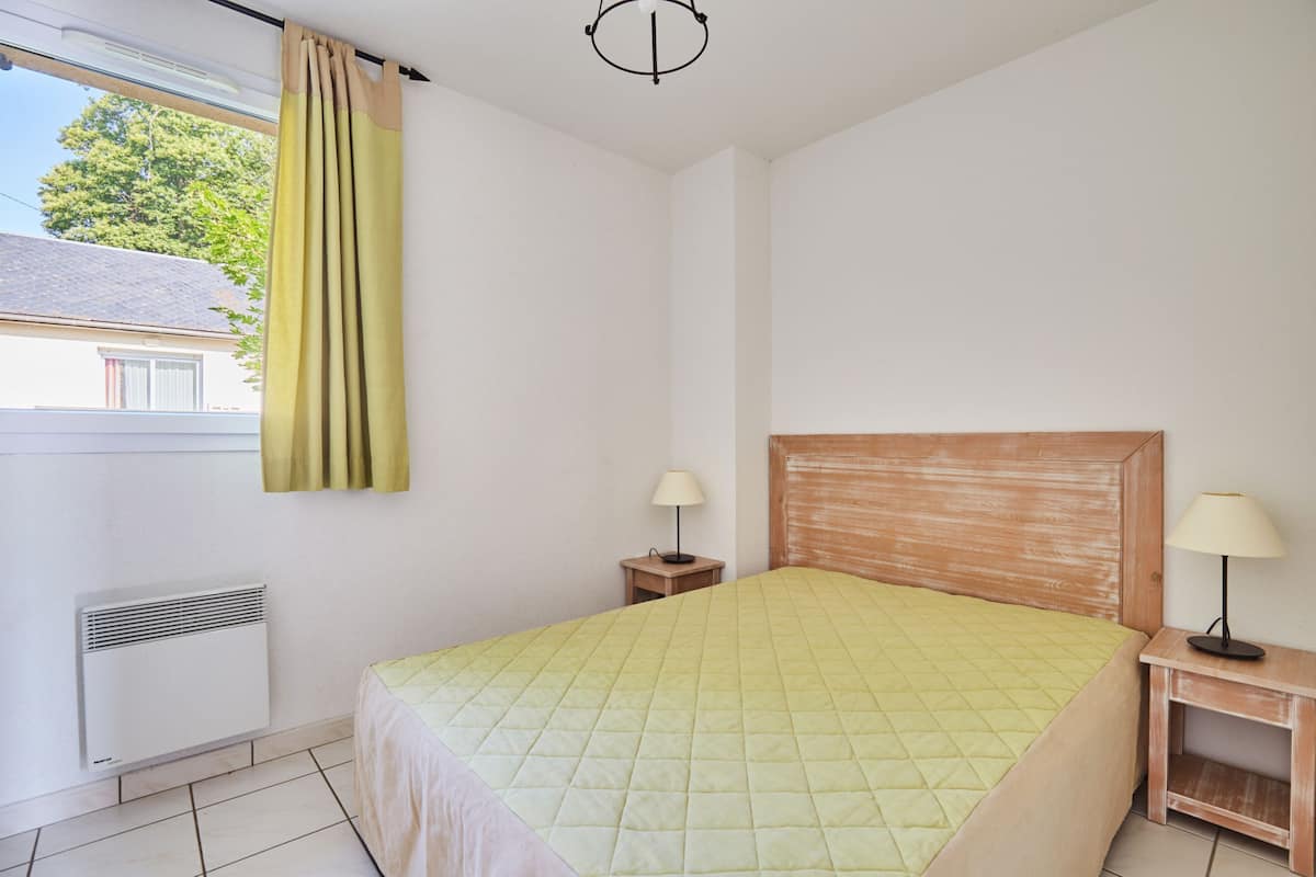 A bedroom with a bed, two bedside tables with lamps, a window with curtains, and a radiator.
