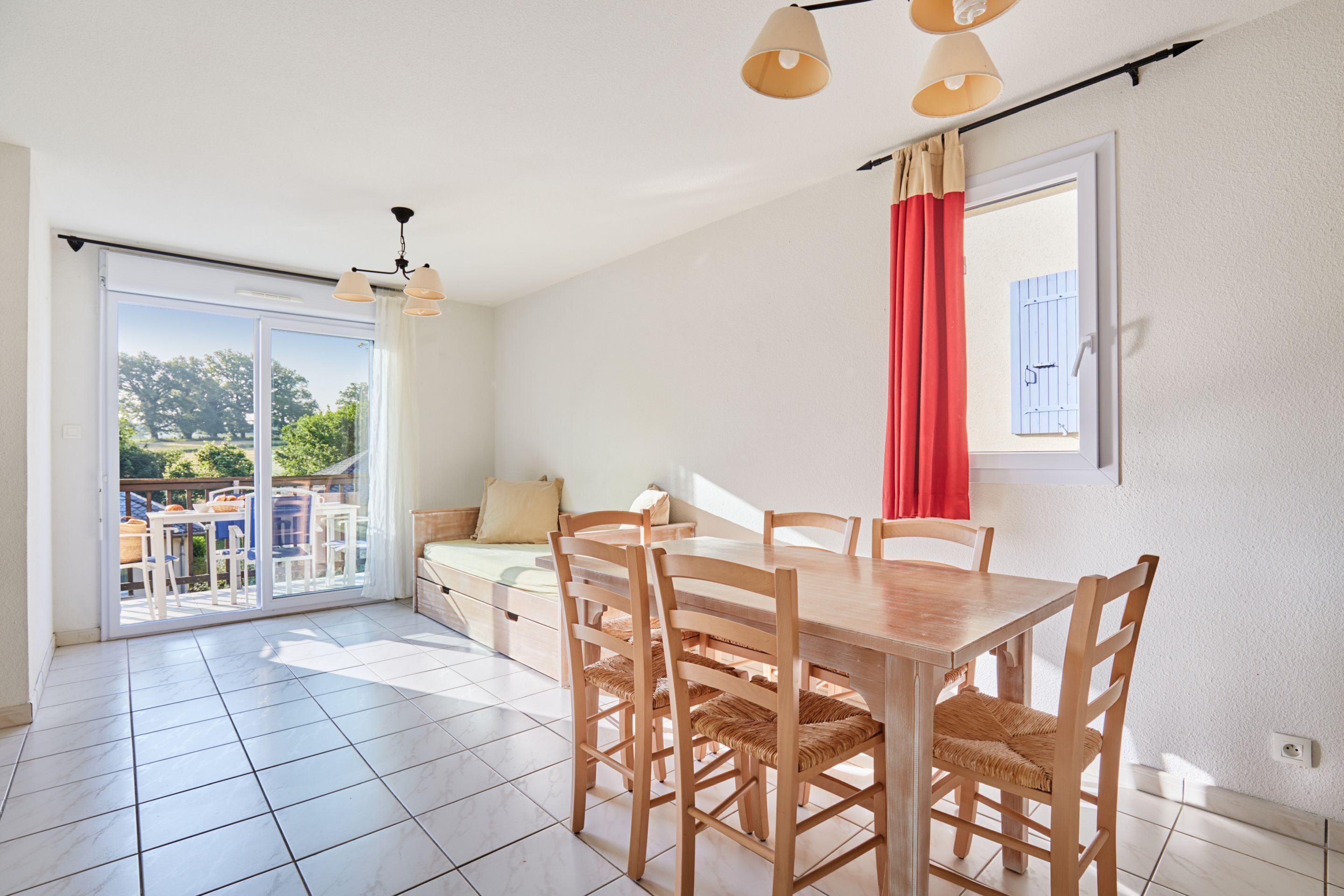 Un coin repas avec une table et des chaises en bois, une porte-fenêtre coulissante donnant sur un espace extérieur aménagé, et un rideau rouge à la fenêtre.
