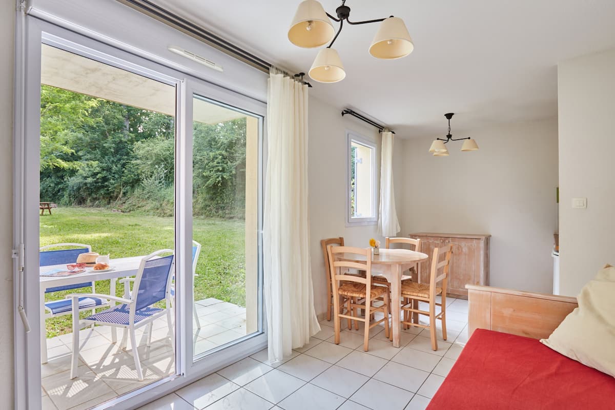 A room with a sliding glass door leading to a garden, a dining area with a table and chairs, and a bed with a red cover.