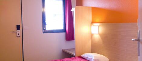 A hotel room with a bed, a window with curtains, a bedside table, and a wall-mounted light.