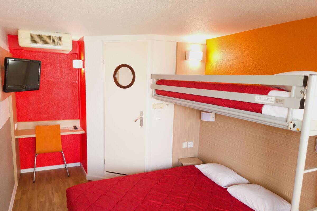 A compact room with a bunk bed, a desk with a chair, a television, and a door with a circular mirror.