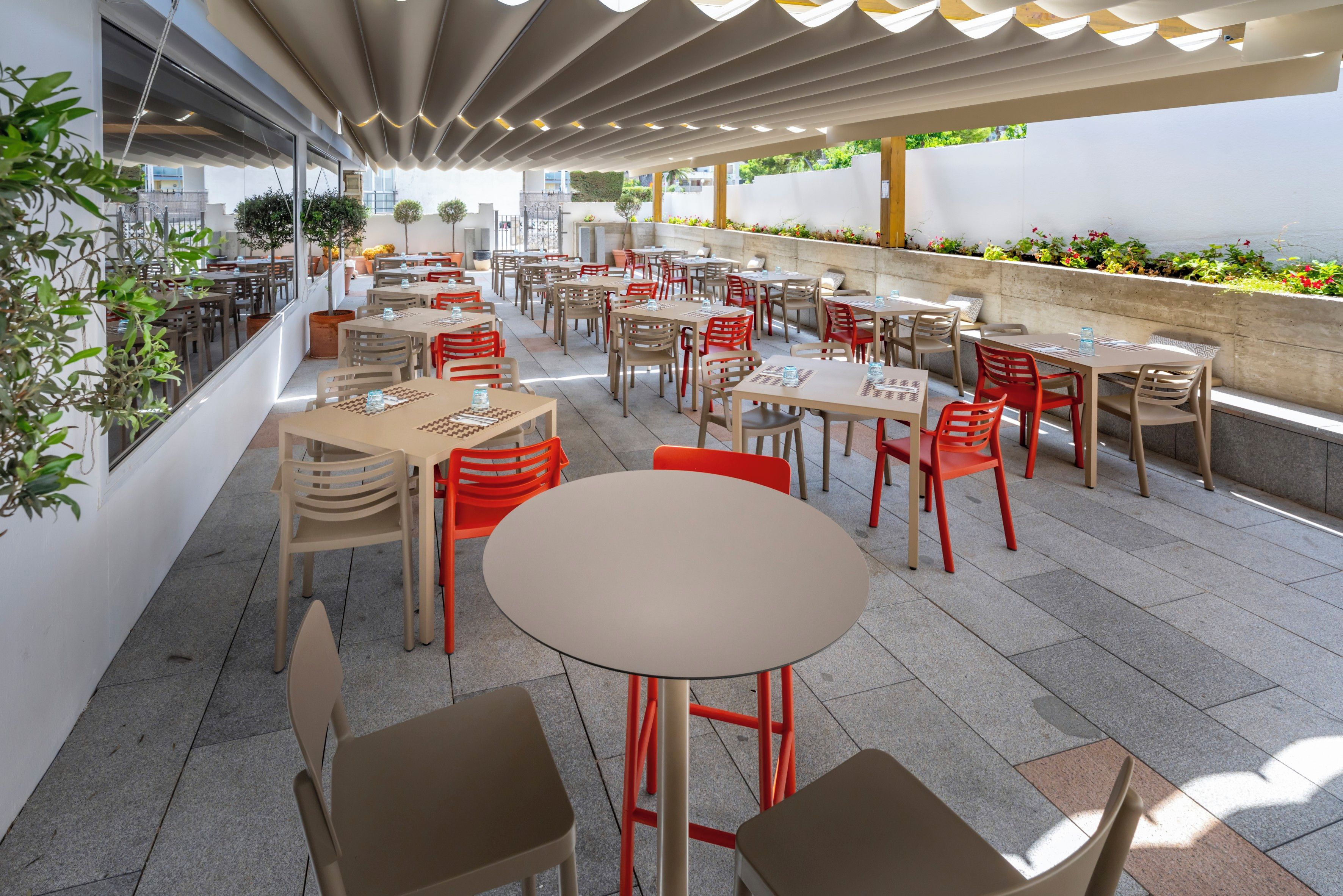 Un café al aire libre con varias mesas y sillas, algunas con detalles en rojo, y una variedad de plantas en macetas.